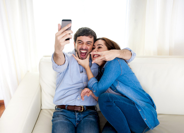 милая привлекательная молодая пара, сидящая вместе на диване, делая селфи фото с мобильного телефона
 - Фото, изображение