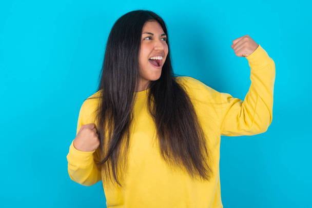 Attractive  latin woman wearing yellow sweatshirt over blue background celebrating a victory punching the air with his fists and a beaming toothy smile - Foto, afbeelding