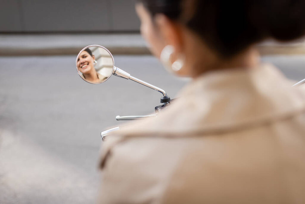 Spiegelbild einer glücklichen Frau im beigen Trenchcoat im Spiegel eines Motorrollers  - Foto, Bild