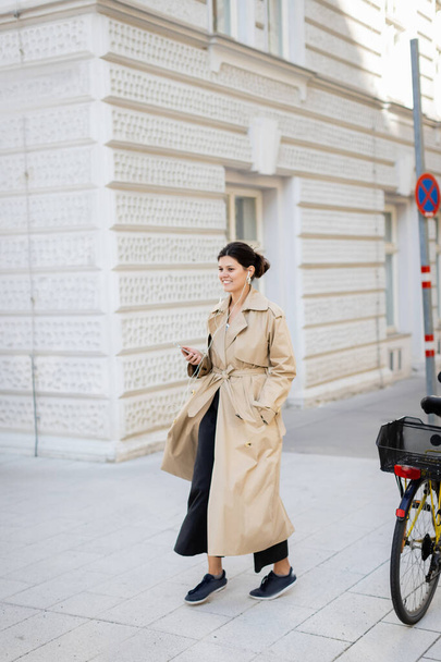 radosna kobieta w słuchawkach trzymająca telefon komórkowy i słuchająca muzyki na ulicy w Wiedniu - Zdjęcie, obraz