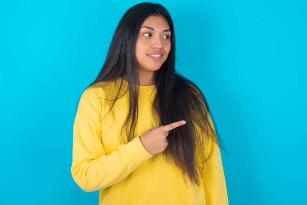 Šťastná veselá usměvavá latinská žena v žluté mikině přes modré pozadí, která vypadá a ukazuje rukou stranou. Kopírovat koncept prostoru a reklamy. - Fotografie, Obrázek