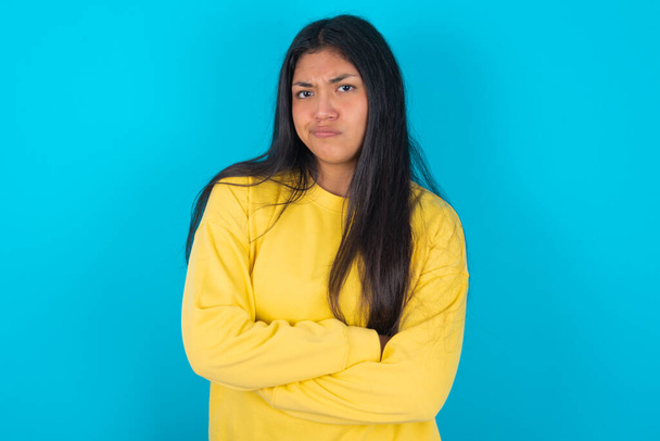 怒っているラテン系女性の写真は,青い背景を横断する腕の上に黄色いスウェットシャツを着ています. がっかりした表情でカメラを見る. - 写真・画像