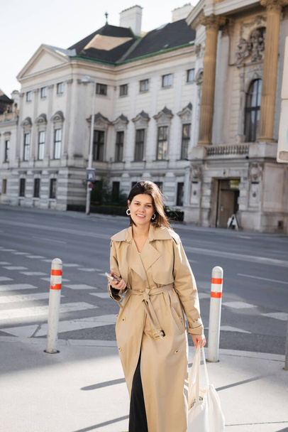 smiling woman in stylish coat holding bag and smartphone on street in vienna - Foto, afbeelding