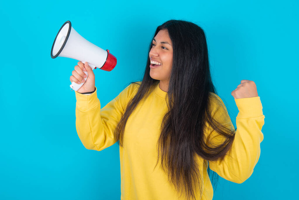 mulher latina vestindo camisola amarela sobre fundo azul comunica gritando alto segurando um megafone, expressando sucesso e conceito positivo, ideia de marketing ou vendas. - Foto, Imagem