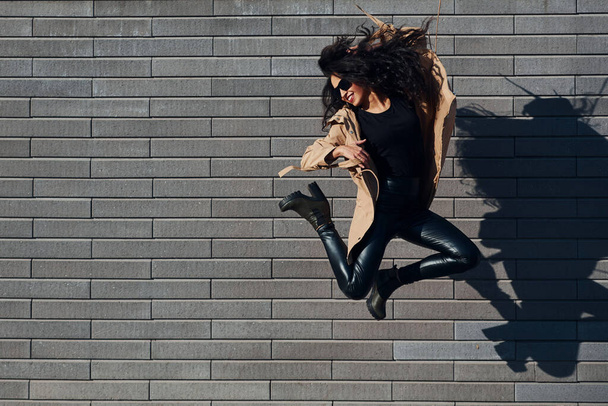 Beautiful brunette with curly hair and in black clothes jumping and having fun outdoors near wall. - Foto, afbeelding