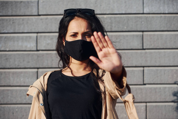 Showing stop gesture by hand. In protective mask. Beautiful brunette with curly hair and in black clothes outdoors near wall. - Фото, зображення