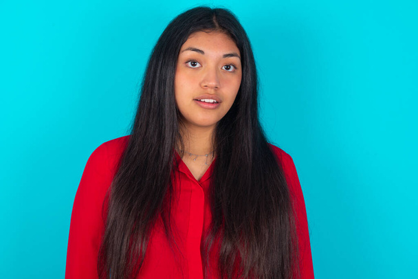 Shocked  latin woman wearing red T-shirt over blue background stares bugged eyes keeps mouth opened has surprised expression. Omg concept - 写真・画像