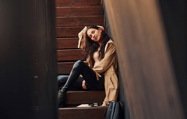 Cheerful brunette in black leggings sitting on wooden stairs and smiling. - 写真・画像