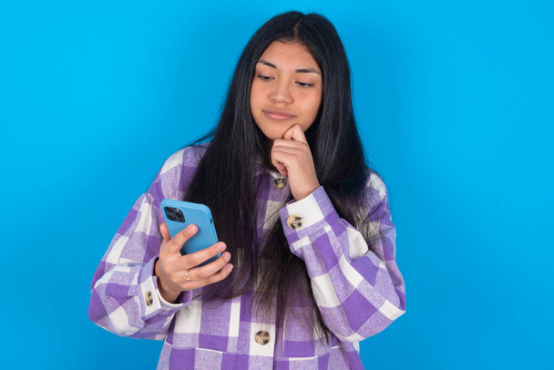 Przemyślana szczęśliwa Latynoska kobieta nosząca koszulę w kratę na niebieskim tle trzymać wygląd telefonu, copyspace - Zdjęcie, obraz