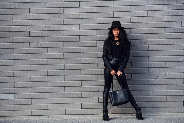 Beautiful brunette with curly hair and in black clothes holding bag outdoors near wall. - Φωτογραφία, εικόνα