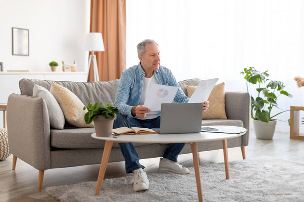 Man holding documents reading report working on laptop at home - Photo, Image