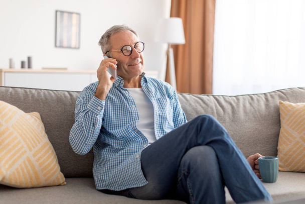Веселый взрослый мужчина разговаривает по мобильному телефону, сидя на диване - Фото, изображение