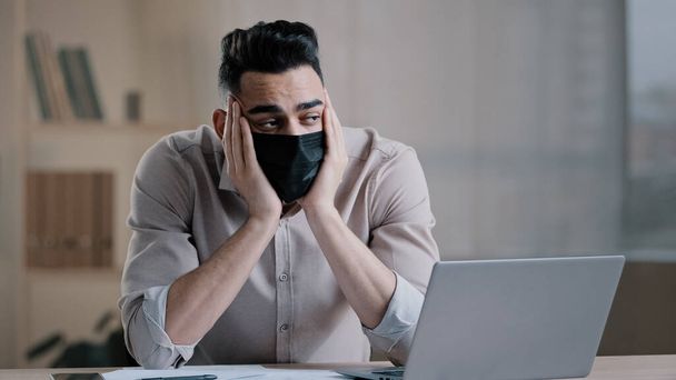 Bored tired hispanic guy worker in medical mask during pandemic coronavirus young business man tired ill worker feel boredom in home office hold head suffering from insomnia thinking about problems - Фото, изображение