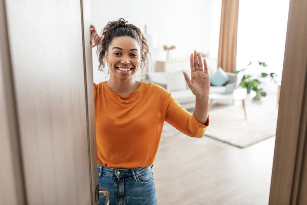 Веселая черная женщина, открывающая дверь дома, машет рукой в помещении - Фото, изображение