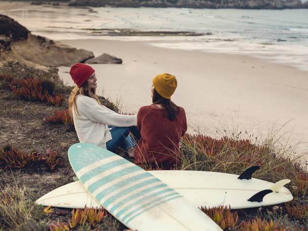 Two best friends sitting near the coastline with her surfboards while looking to the ocean - Photo, image