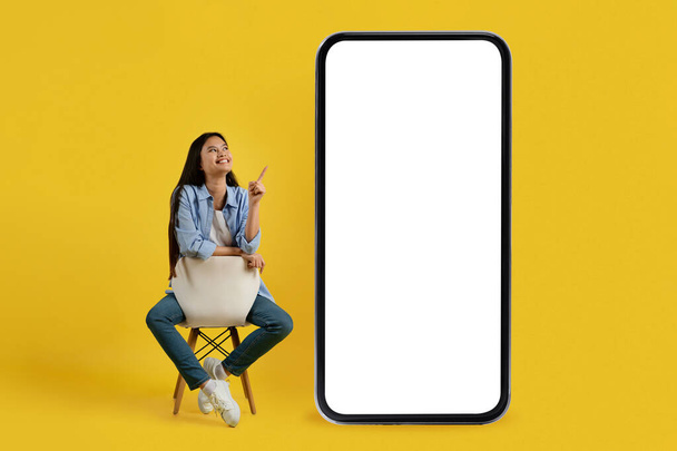 Feliz joven japonesa hembra en casual sentado en la silla y señalando el dedo en el teléfono inteligente enorme con la pantalla vacía - Foto, imagen