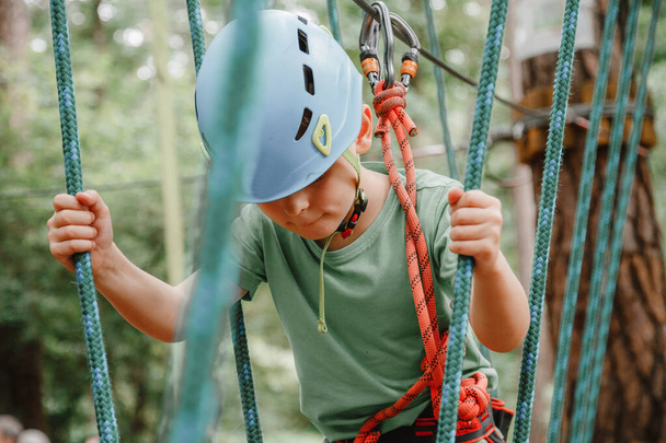 Brave little boy having fun at adventure park and smiling to camera wearing helmet. Scout practicing rappelling. Child - a boy and a girl in the rope park pass obstacles. - Фото, изображение