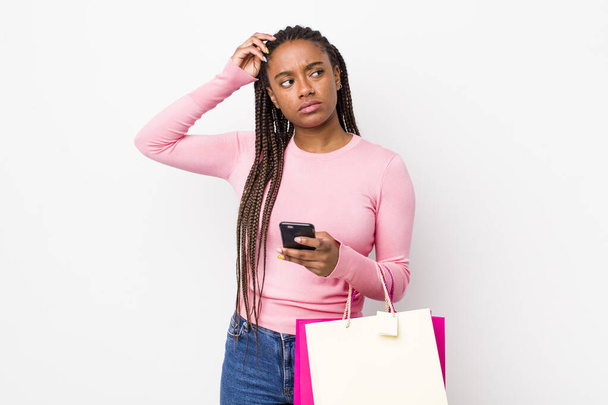 young adult black woman feeling puzzled and confused, scratching head. shopping bags concept - Fotó, kép