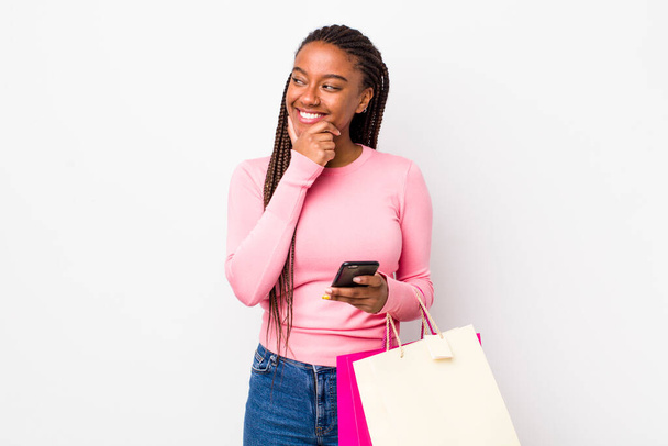 young adult black woman smiling with a happy, confident expression with hand on chin. shopping bags concept - 写真・画像