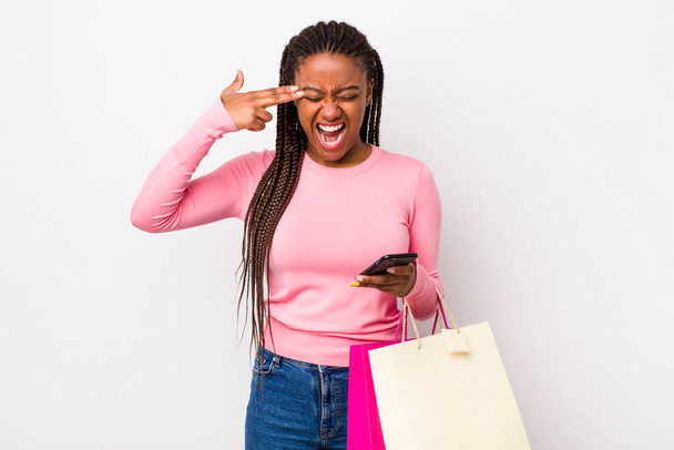 young adult black woman looking unhappy and stressed, suicide gesture making gun sign. shopping bags concept - Foto, Imagen