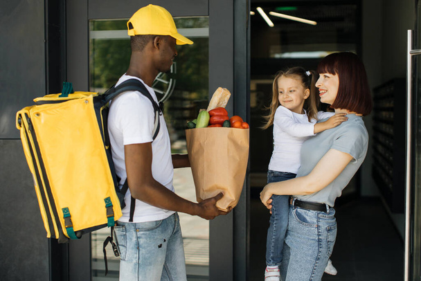 African American delivery man in yellow uniform delivering paper eco bag with grocery takeout food - Φωτογραφία, εικόνα