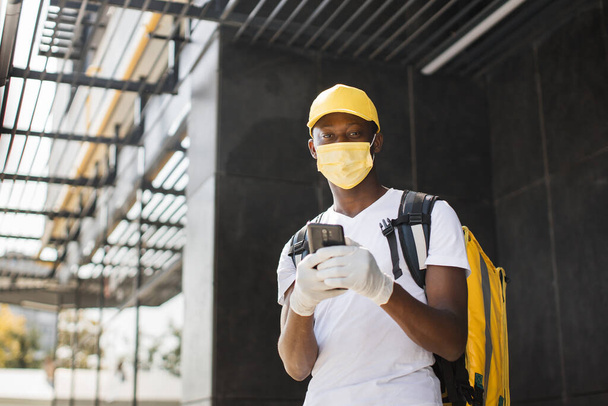 African American male courier food delivery in face mask with yellow thermal backpack - Foto, afbeelding
