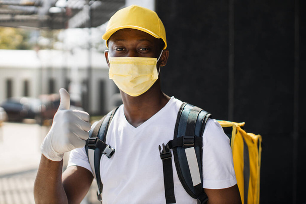 Portrait of delivery man with yellow cap in face mask and thermal backpack looking at camera. - Photo, image