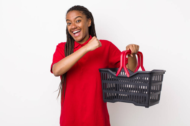 young adult black woman feeling happy and facing a challenge or celebrating. empty shopping basket concept - Valokuva, kuva