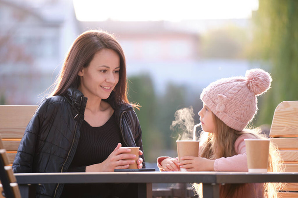 Молодая красивая женщина хорошо проводит время со своей дочерью, сидя в уличном кафе с горячими напитками в солнечный осенний день. Счастье в семейных отношениях - Фото, изображение