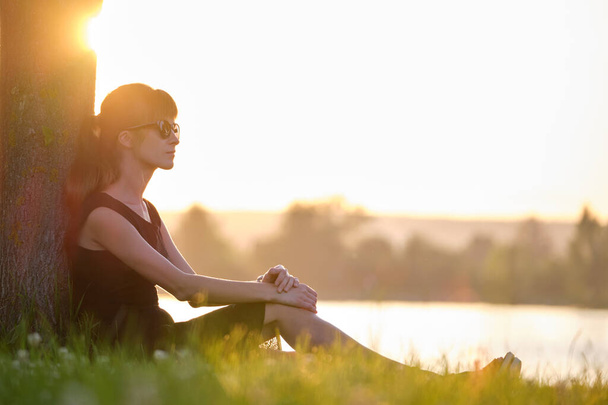 Jeune femme en tenue décontractée assise sur la pelouse verte se détendre sur le bord du lac le soir chaud. Vacances d'été et concept de voyage - Photo, image