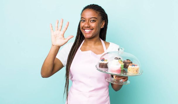 young adult black woman smiling and looking friendly, showing number five. home made cakes concept - Фото, зображення
