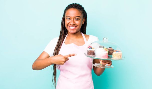 young adult black woman smiling cheerfully, feeling happy and pointing to the side. home made cakes concept - Foto, Imagem