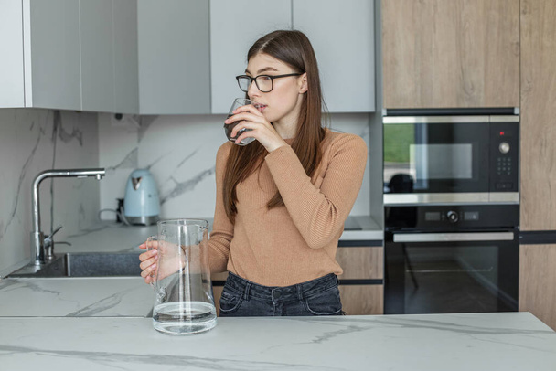 Clean water in glass and in carafe. Young woman in eyeglasses drinking water in modern kitchen. - Photo, Image