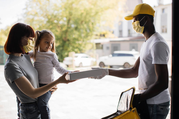 African American delivery man in yellow face mask, uniform delivering parcel box to recipient - Фото, изображение