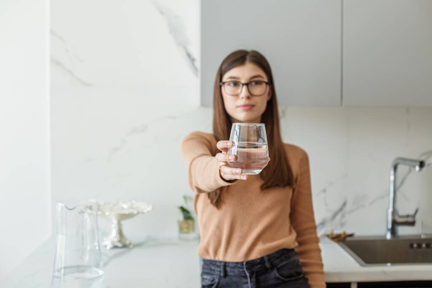 Clean water in glass and in carafe. Young woman in eyeglasses drinking water in modern kitchen. Copy space. - Foto, imagen