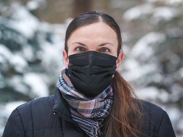 Jonge vrouw in winterjas met zwart wegwerp eenmalig gebruik gezicht virus masker, wazig sneeuw bedekte bomen achtergrond - Foto, afbeelding