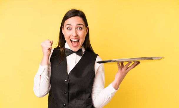 young adult pretty woman feeling shocked,laughing and celebrating success. waiter and tray concept - Foto, imagen