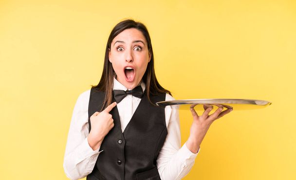 young adult pretty woman looking shocked and surprised with mouth wide open, pointing to self. waiter and tray concept - Zdjęcie, obraz