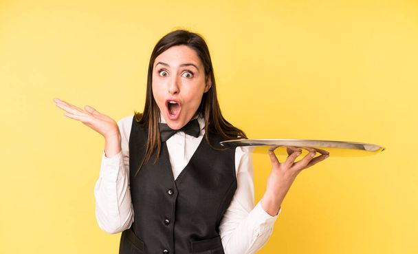 young adult pretty woman looking surprised and shocked, with jaw dropped holding an object. waiter and tray concept - Zdjęcie, obraz