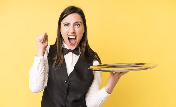 young adult pretty woman shouting aggressively with an angry expression. waiter and tray concept - Valokuva, kuva