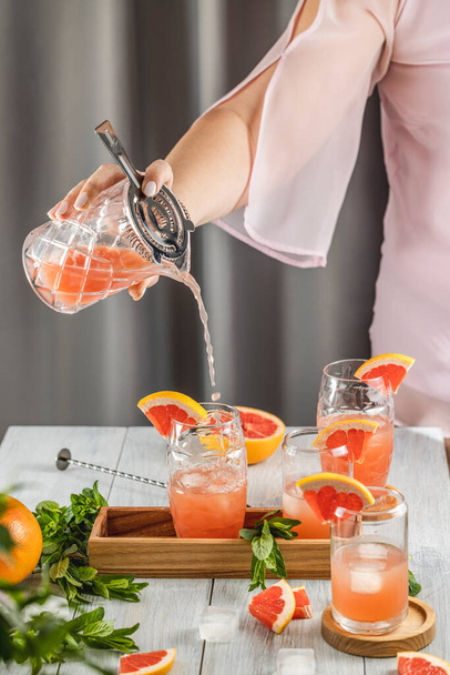 Real woman preparing Grapefruit Cocktail in highball glass or mocktails surrounded by ingredients and bar tools on light gray table surface. Ready for drinking - Foto, immagini