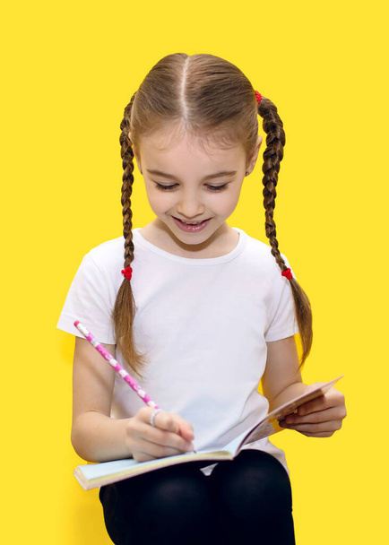 Mädchen mit niedlichen Zöpfen lernen schreiben oder zeichnen, sitzen auf gelbem Hintergrund und lächeln. Das Kind hält ein Notizbuch in der Hand und schreibt mit einem Bleistift Hausaufgaben - Foto, Bild