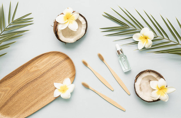 Bambus-Zahnbürste und ätherisches Öl auf einem Tisch mit Kopierraum auf weißem Hintergrund. Gestylte Komposition aus flacher Lage mit Kokosnuss, tropischen Blättern und Blüten. - Foto, Bild