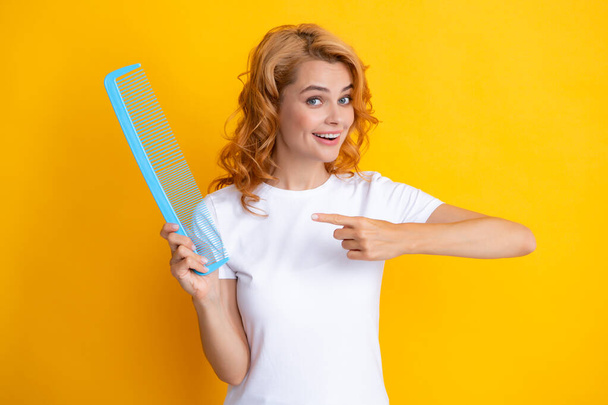 Young woman hairdresser with comb. Funny portrait of redhead woman pointing on comb, isolated on yellow background. - Фото, изображение