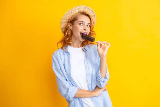 Молодая женщина ест мороженое с шоколадной глазурью на желтом фоне. Смешная рыжая девчонка в летней соломенной шляпе лижет мороженое.. - Фото, изображение