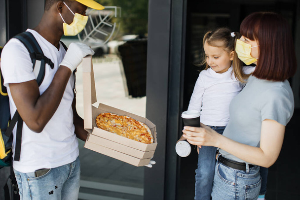 African American delivery man in yellow face mask, uniform cap delivering cardboard boxes with pizza - Foto, afbeelding