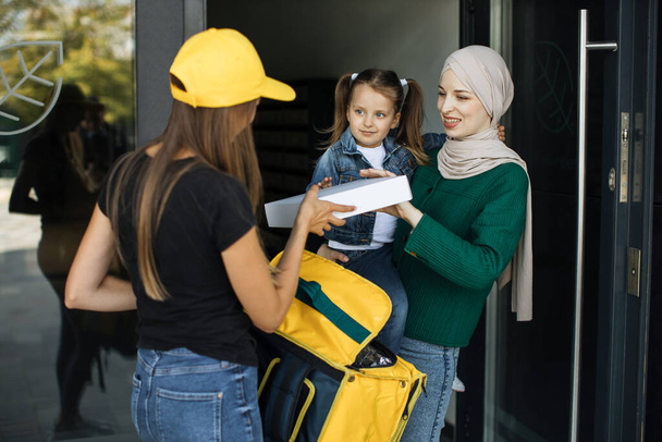 Beautiful young islamic woman with her cute girl opening the door of her house and meets courier - Фото, изображение