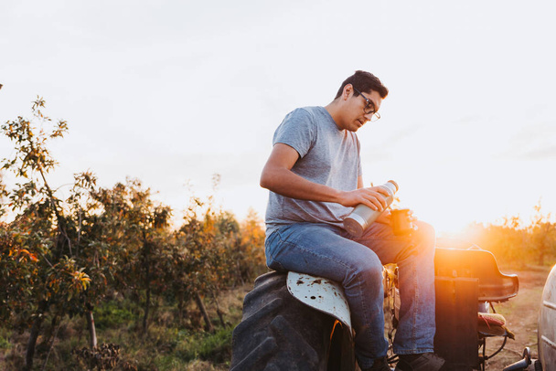Young latin man drinking mate and relaxing, in a rural space, on top of an old little tractor. Hot beverage - Fotografie, Obrázek