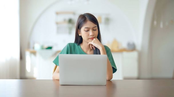 Ασιάτισσα νεαρή γυναίκα εργάζεται σοβαρά στον υπολογιστή του σπιτιού. Σκέφτεται να βρει λύση στο πρόβλημα της εργασίας - Φωτογραφία, εικόνα