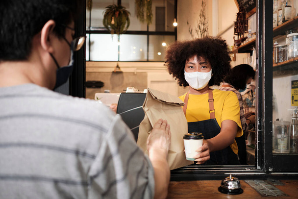 African American γυναίκα barista με μάσκα προσώπου λειτουργεί στο καφέ με κοινωνική απόσταση, takeaway καφέ για έναν πελάτη, νέα κανονική υπηρεσία των μικρών επιχειρήσεων καφέ στο COVID19 καραντίνα τρόπο ζωής. - Φωτογραφία, εικόνα